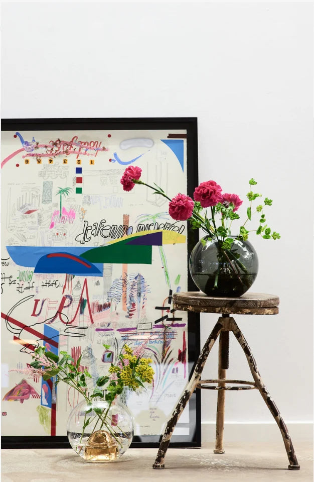 Black glass vase of red flowers on a twig stool in front of an abstract painting