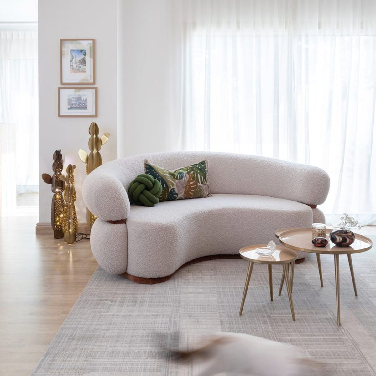 Malibu Curved Sofa in cream with green pillows beside gold nesting tables