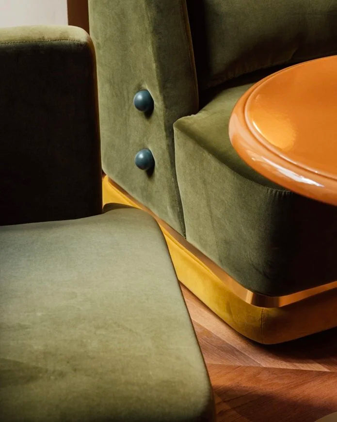 Panorama Armchair in olive velvet with brass base and dark buttons beside an amber side table