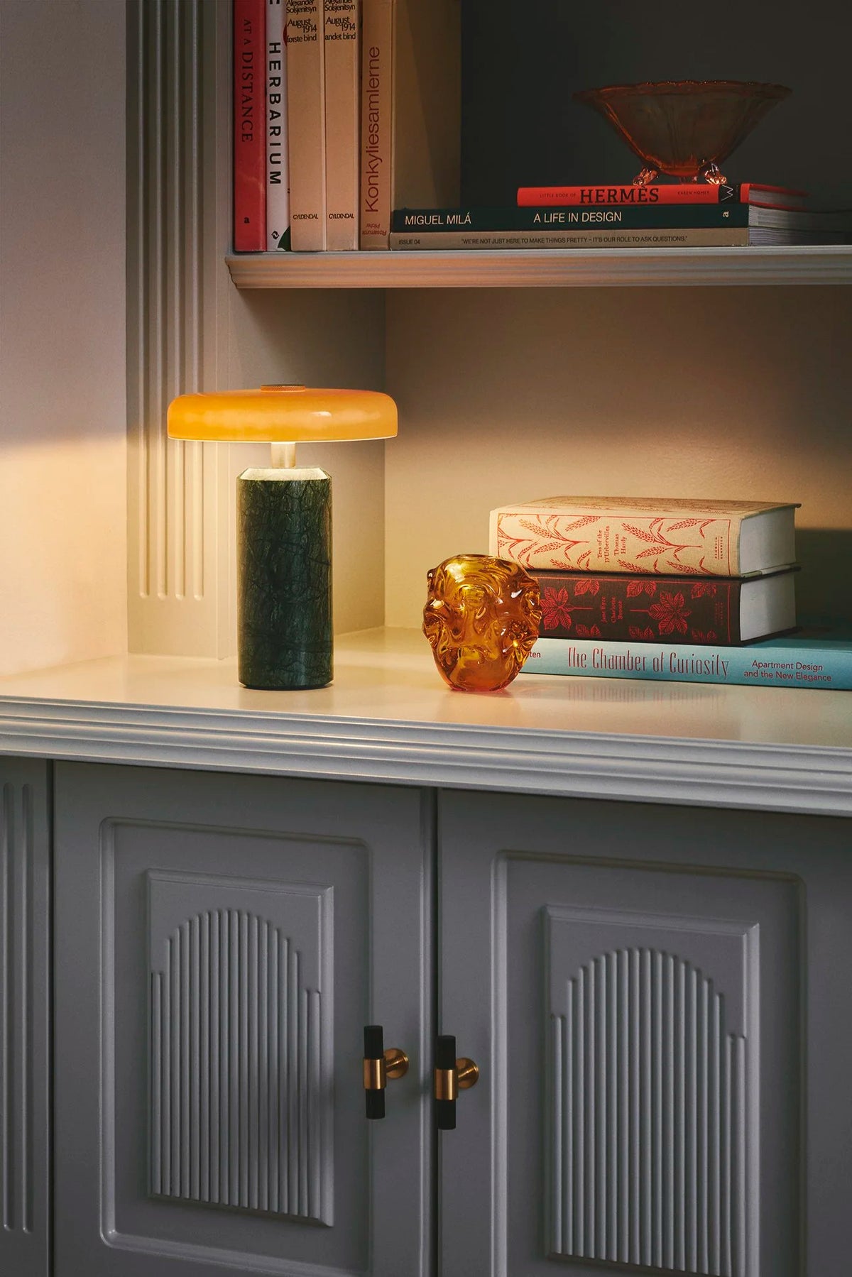 Trip Table Lamp in Moss and Amber glowing on a shelf with books