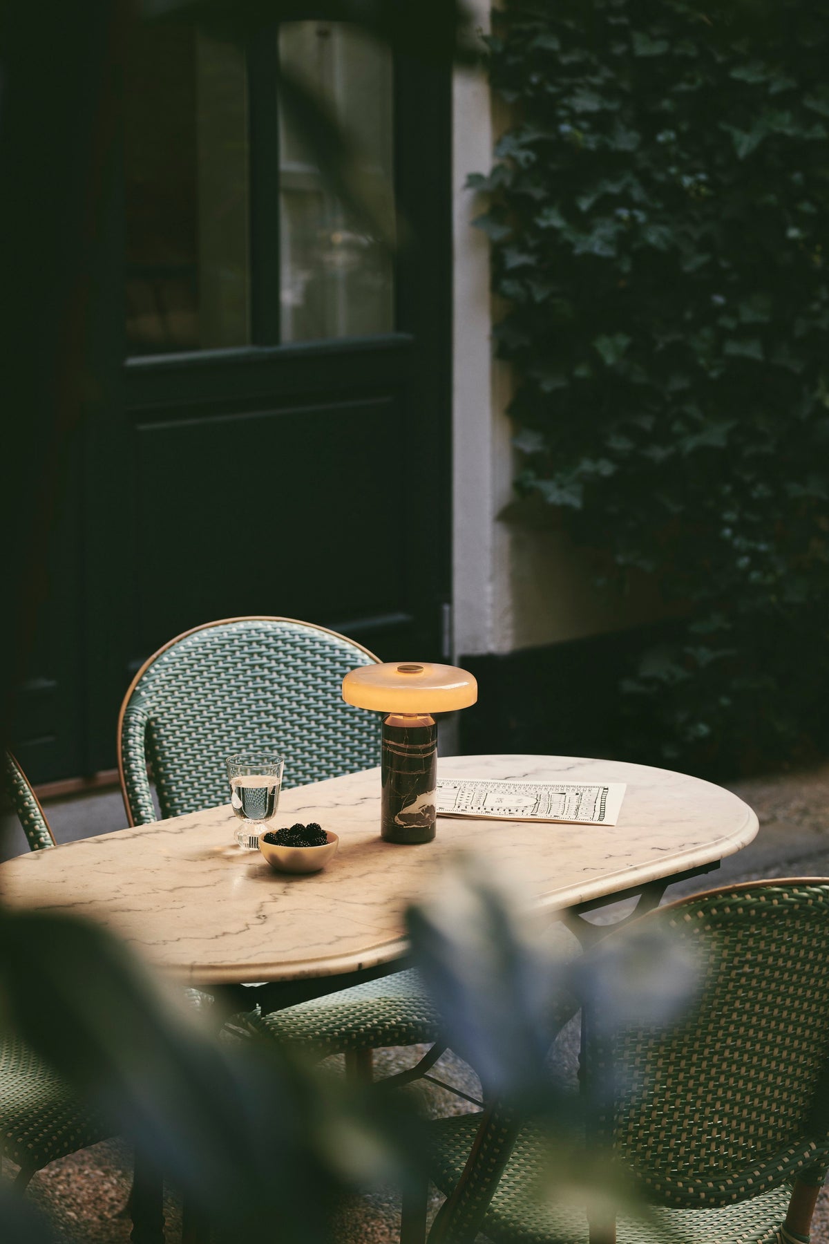 Trip Table Lamp glowing on a marble café table beside green rattan chairs