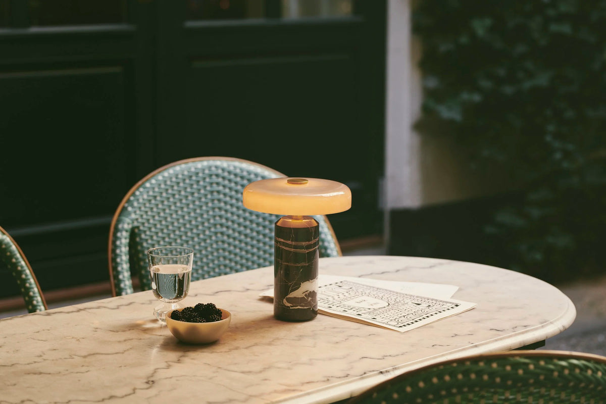 Trip Table Lamp – Ash | Grey on a café table with rattan chairs, wide view