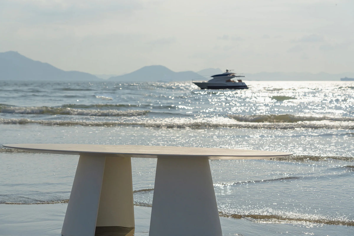 Tumuli Oval Dining Table in white on the shoreline with a boat in the distance