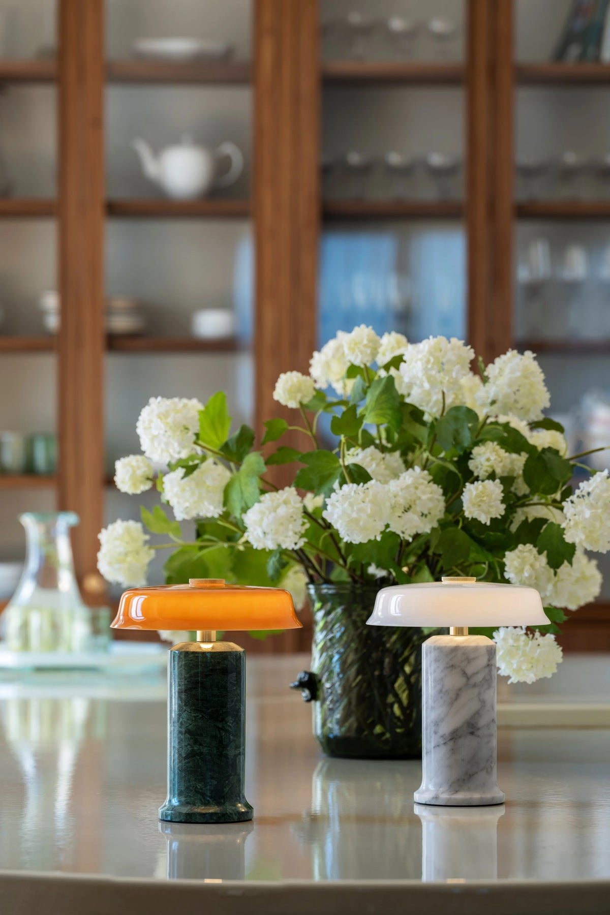 Trip 2 Table Lamp in Moss and Amber beside a white version and flowers on a counter