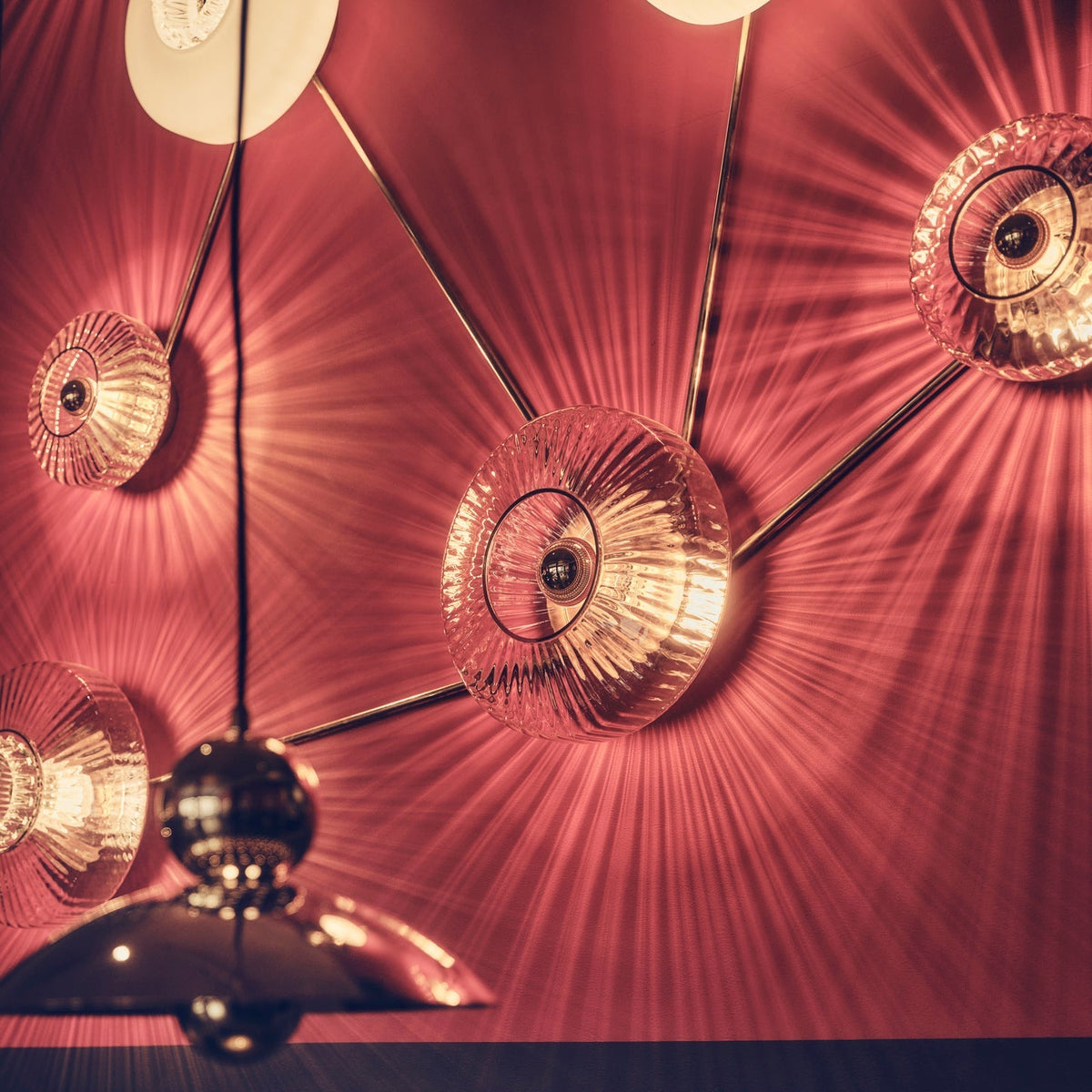 New Wave Optic Wall Sconce XL lights glowing on a deep red wall beside a brass chandelier