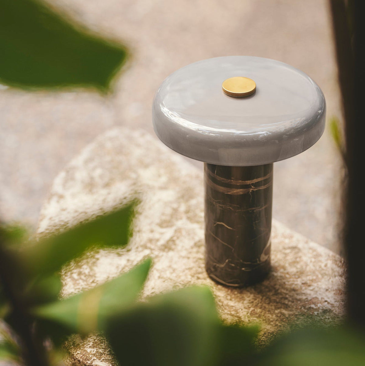 Trip Table Lamp – Ash | Grey switched off on a stone among green leaves