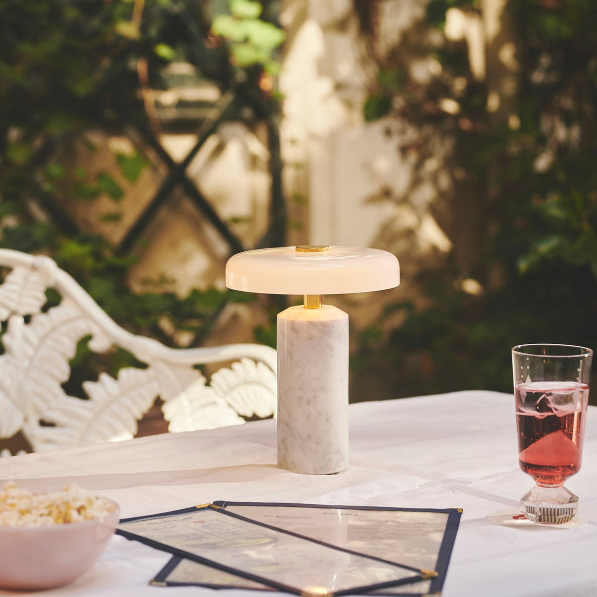 Trip Table Lamp in Carrara / Opal lit on a garden table with white iron bench