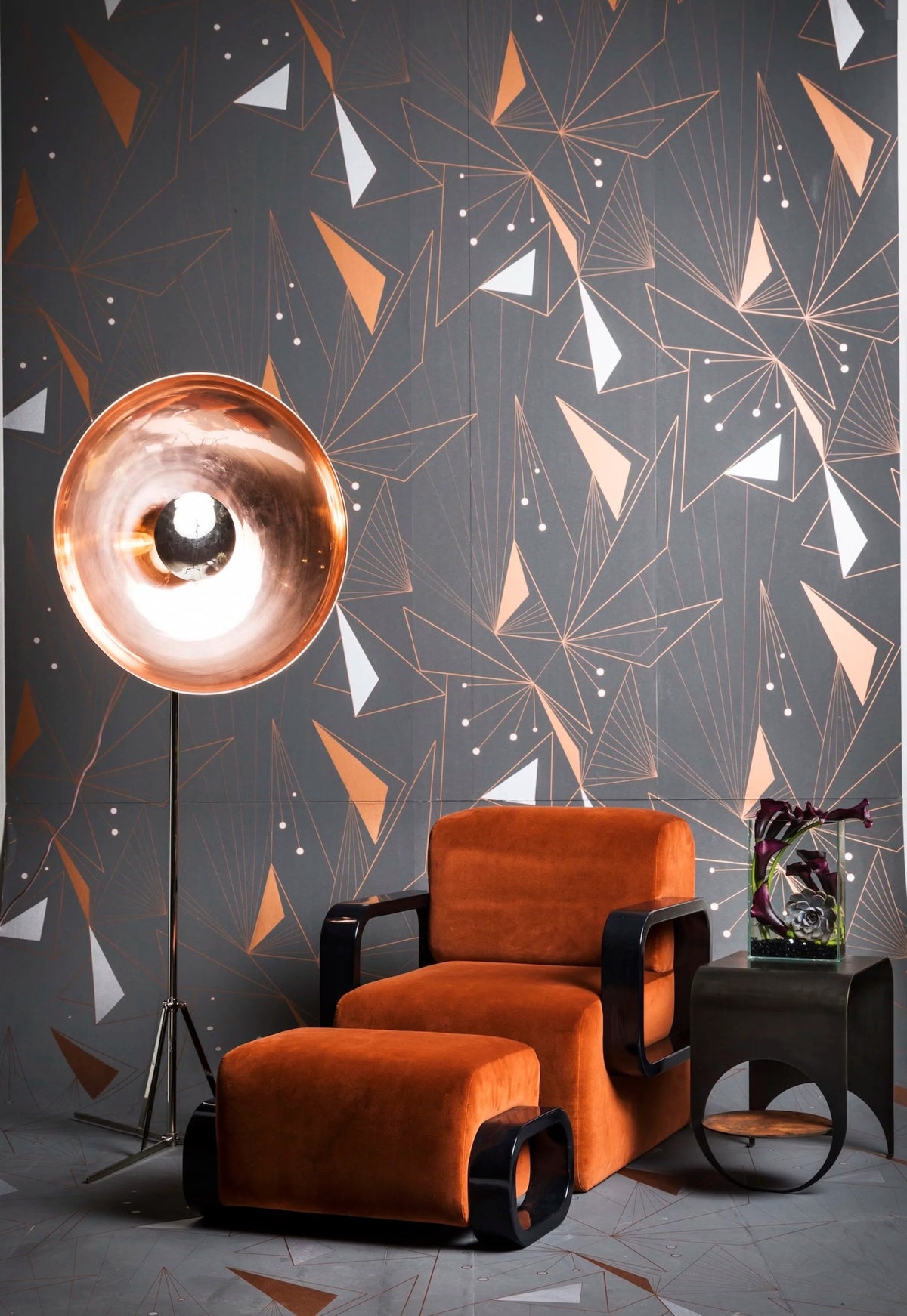 Orange velvet armchair and ottoman against a geometric black wall with a copper floor lamp