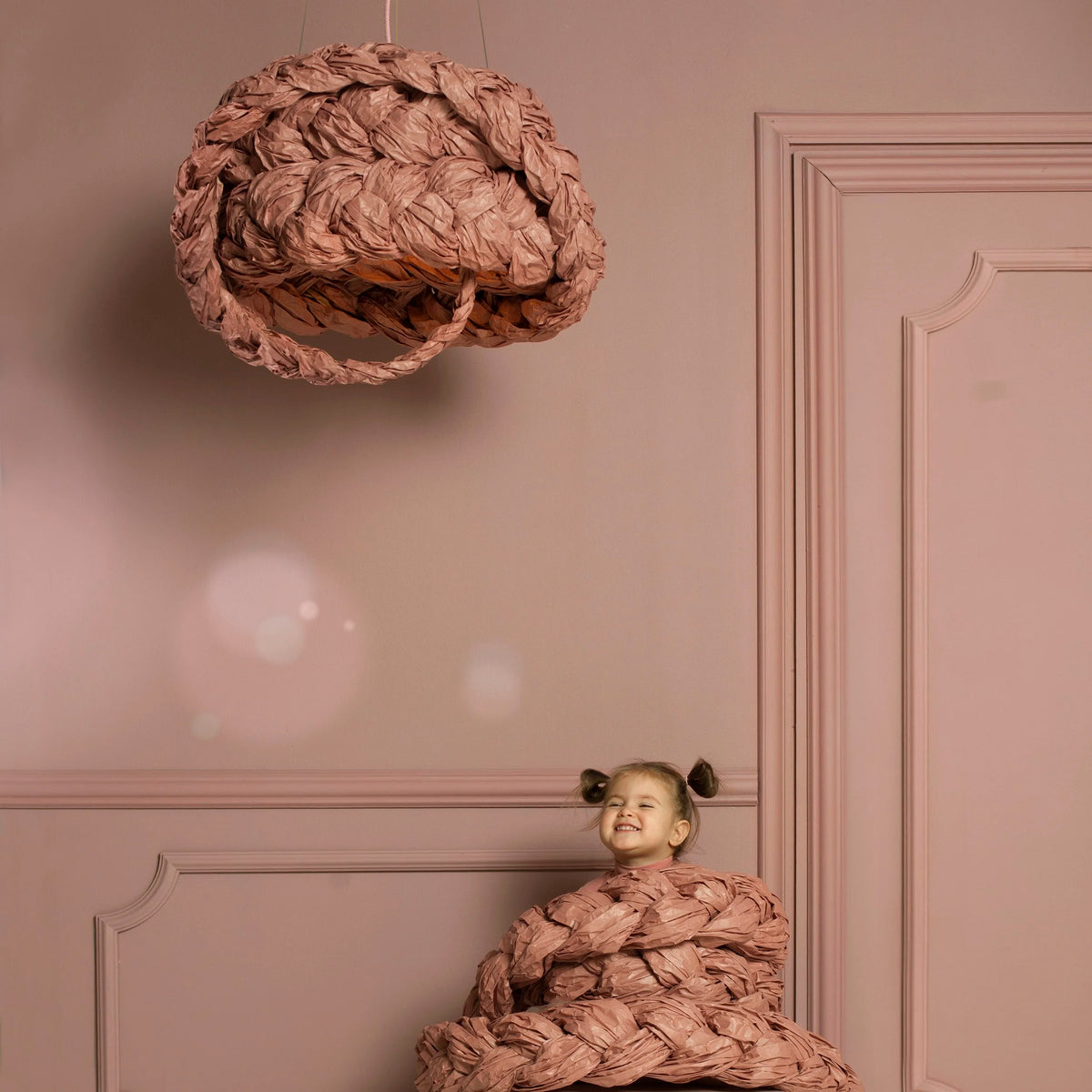 Mammalampa Bride Pendant Light in pink above a smiling child wrapped in braided pink cord