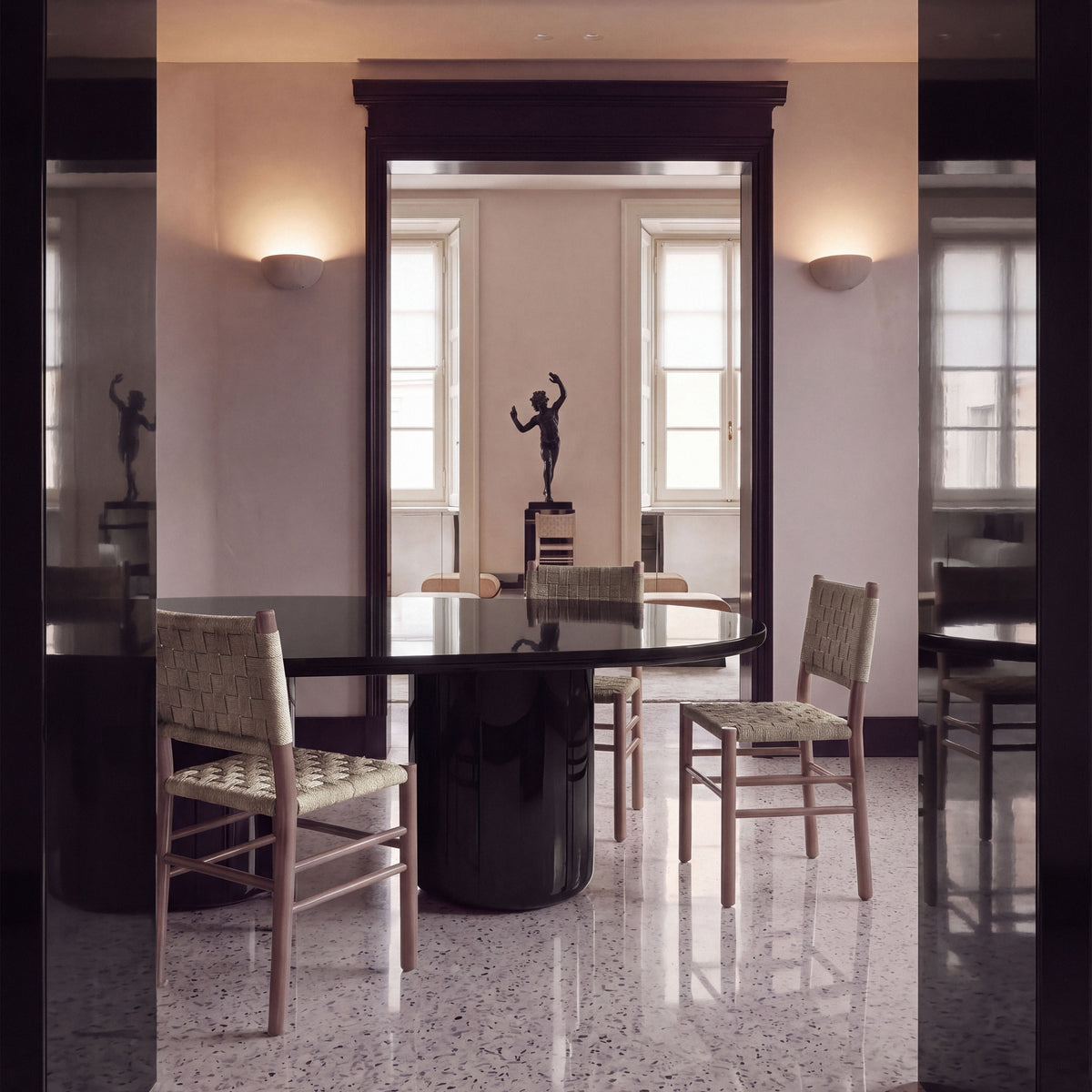 Laghi II Dining Table in black lacquer with rush chairs below wall sconces and a statue