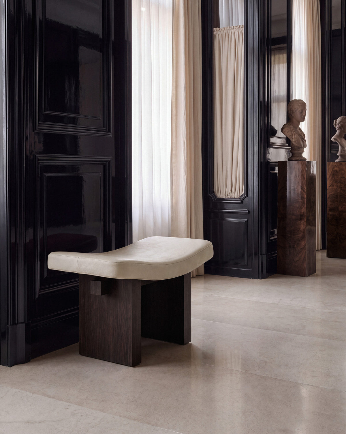 Nara Ottoman in cream on dark wood on a stone floor beside a sculpture bust