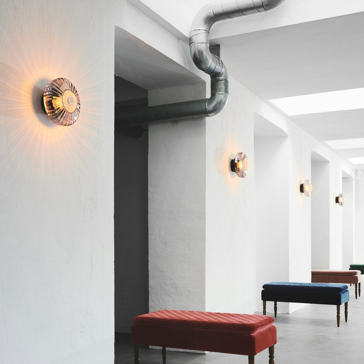 New Wave Optic Wall Sconce series lit along a white industrial corridor with upholstered benches