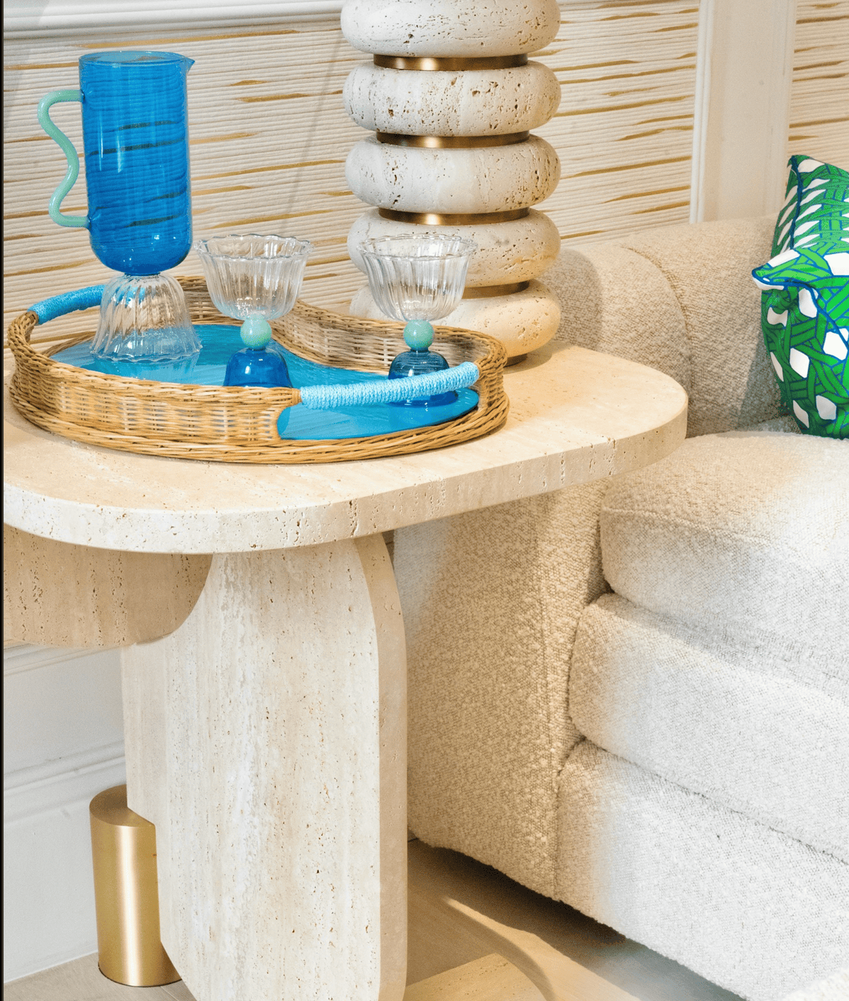 Playing Games Travertine Side Table beside a bouclé sofa with a tray of blue glassware