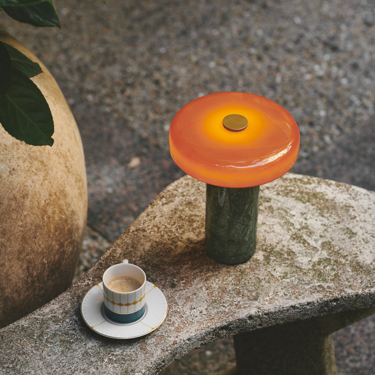 Trip Table Lamp in Moss / Amber lit on a stone garden bench beside a coffee cup